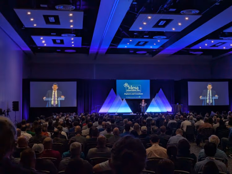 A speaker at the podium in front of a packed ballroom at Clean Currents 2026, shown on two large side screens